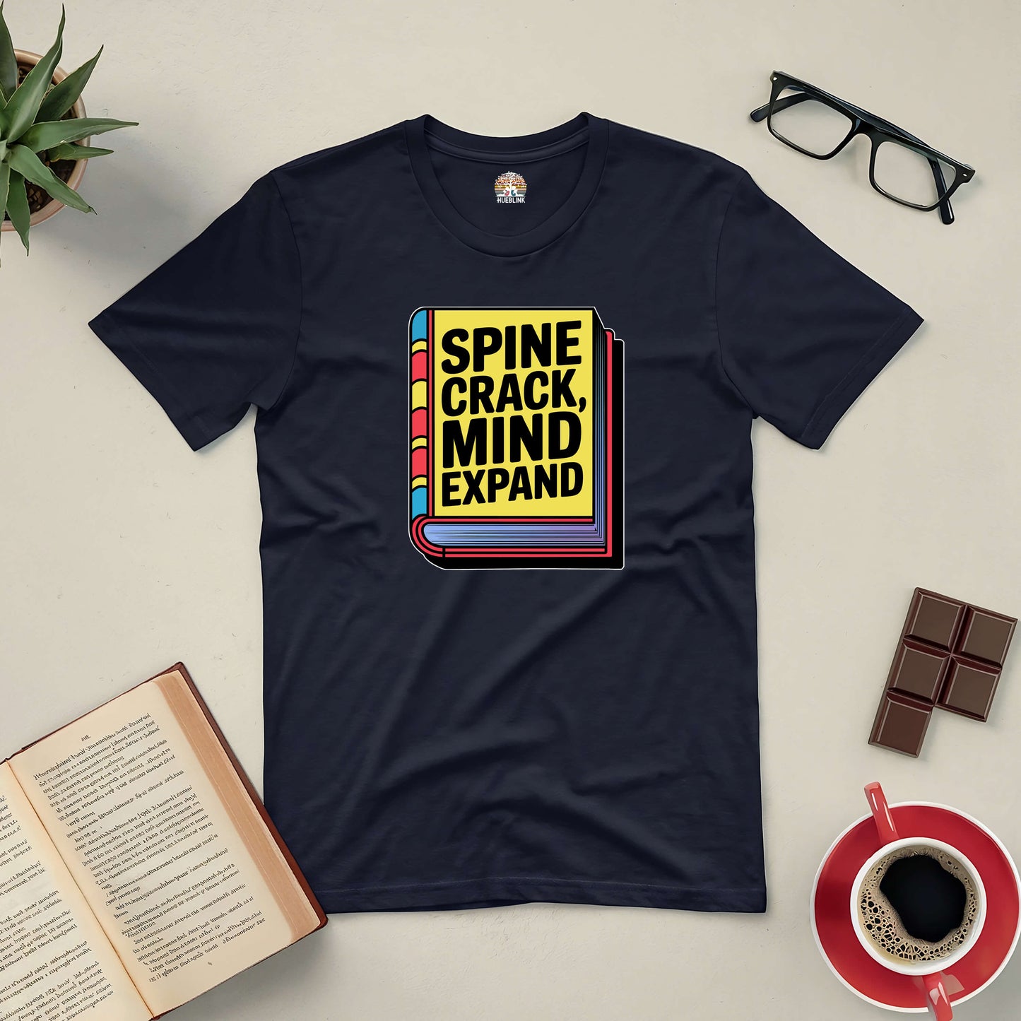 "Spine Crack, Mind Expand Tee with book graphic, surrounded by an open book, glasses, chocolate, coffee, and a plant"