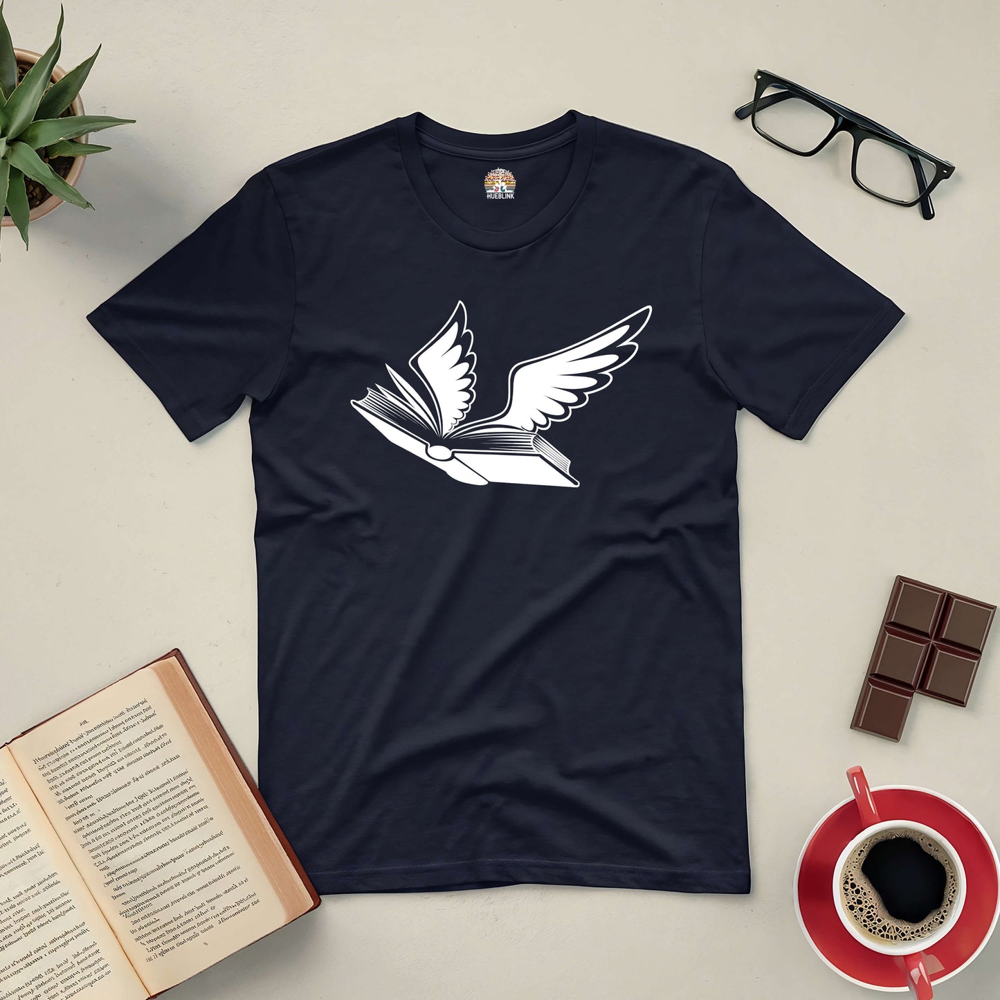 Black Bird of Hermes tee with winged book design, surrounded by open book, glasses, succulent, chocolates, and coffee cup.