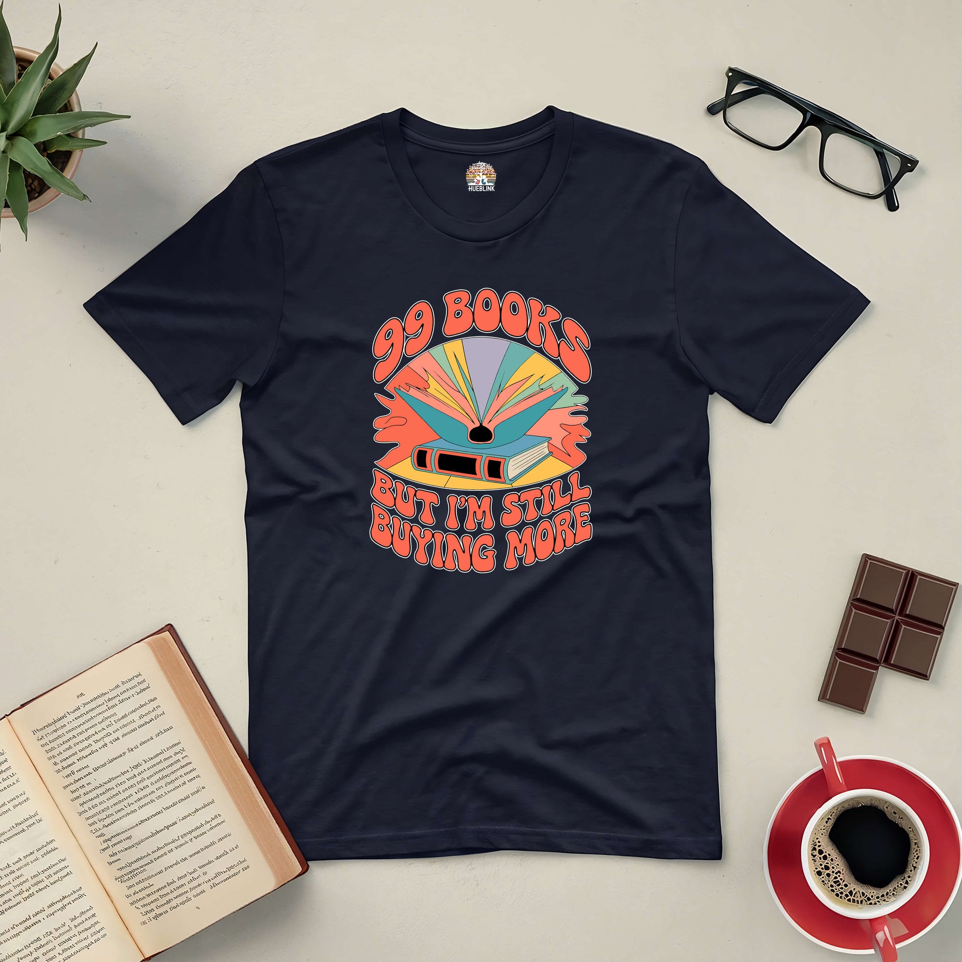 Black "99 Books and Counting!" tee on a table with open books, coffee, and glasses, showcasing a passion for reading.