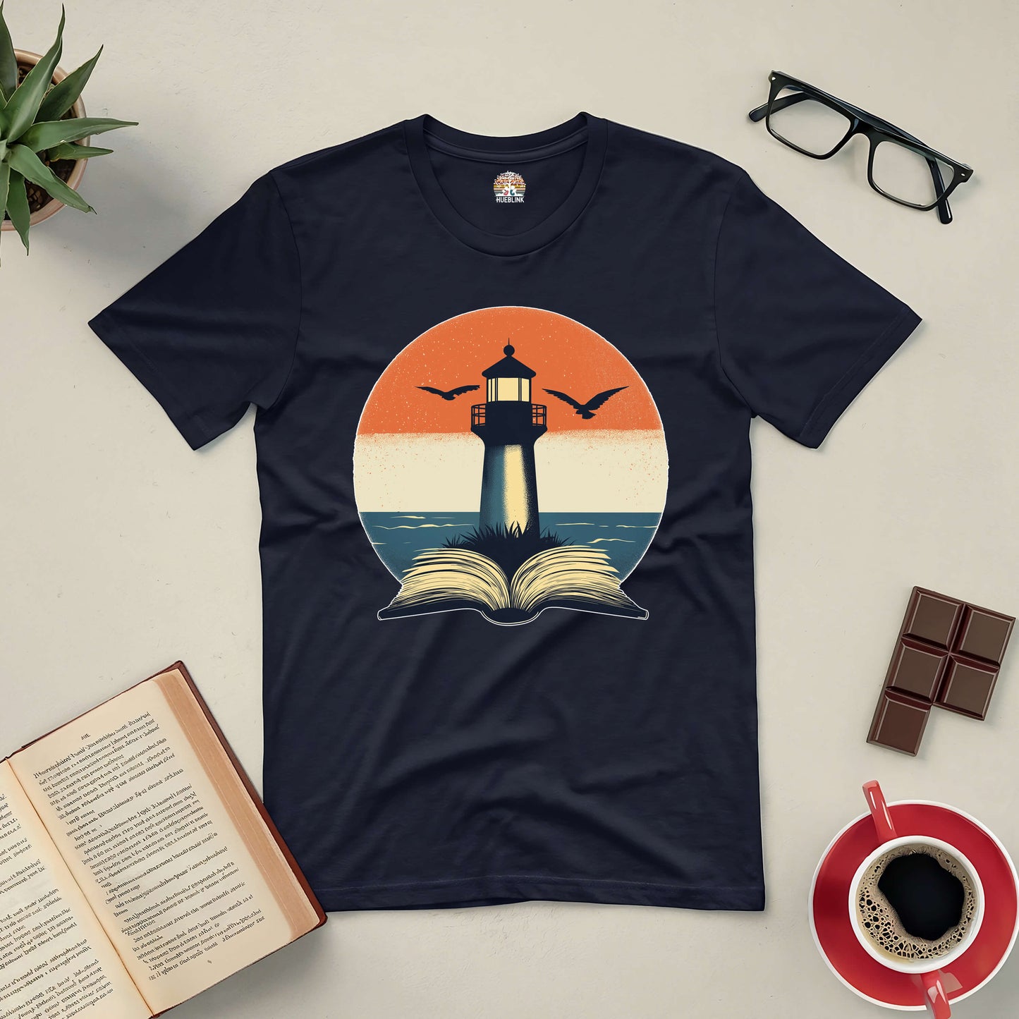 Black t-shirt featuring a lighthouse and open book design, placed on a table with glasses, coffee, a plant, chocolate, and an open book.