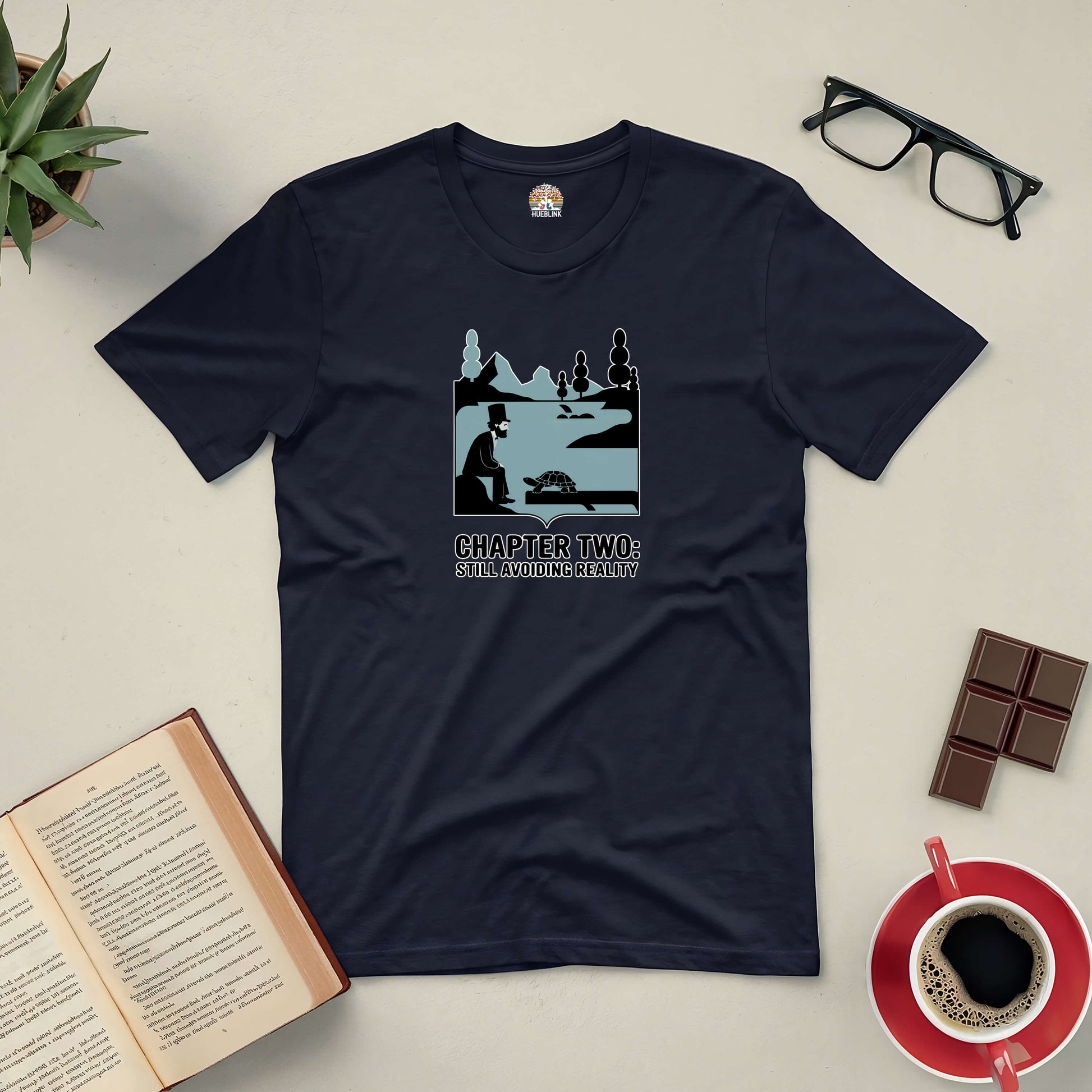 "Chapter Two: Still Avoiding Reality Tee with book, coffee, glasses, and chocolate on a table setting"