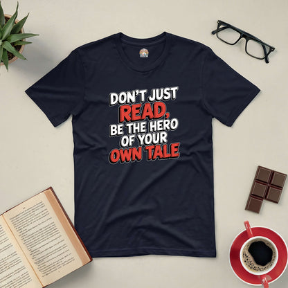 "Navy blue tee with 'Don't Just Read, Be the Hero of Your Own Tale' text on a flat lay with book, coffee, chocolate, and glasses."