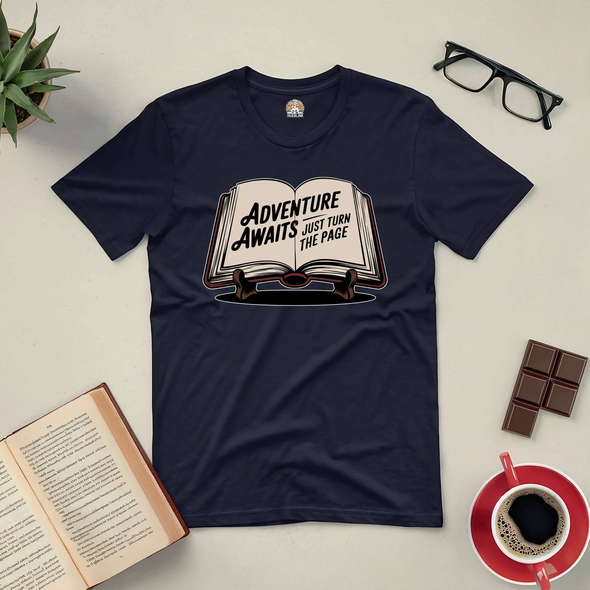 "Adventure Awaits tee with book design, surrounded by glasses, a book, chocolate, coffee, and a plant on a table"