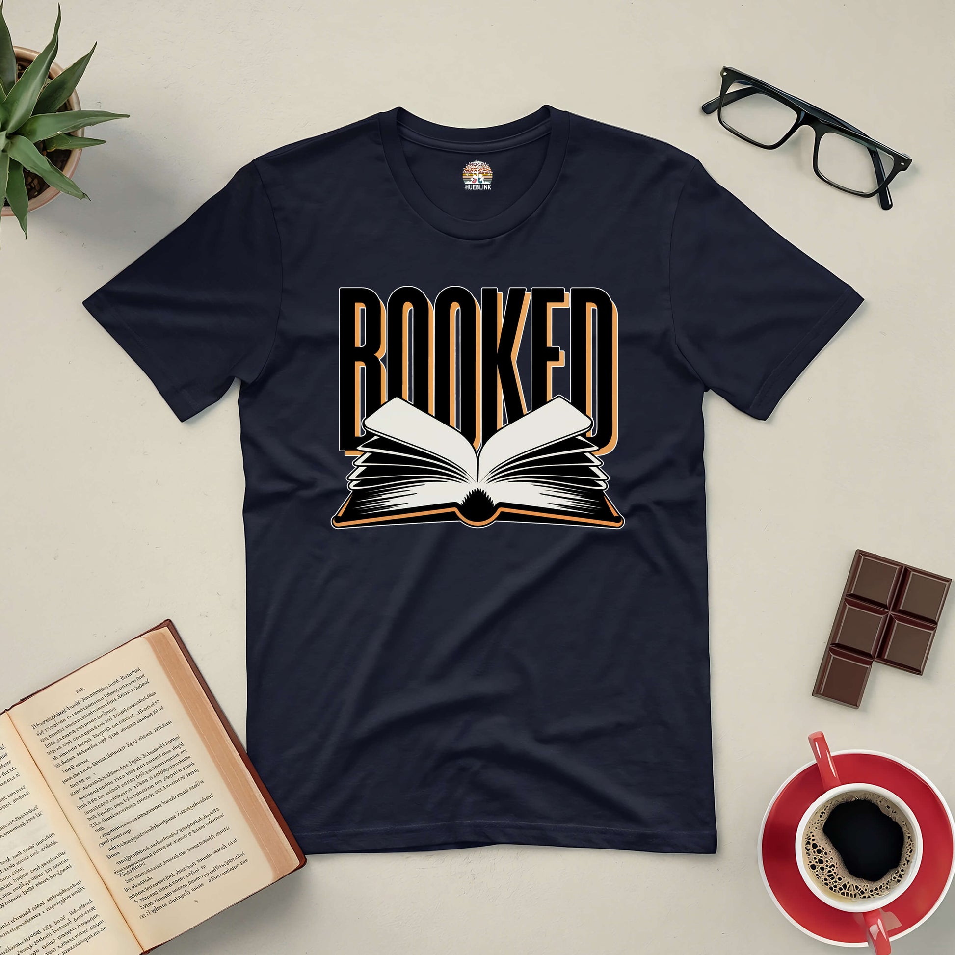 "Booked Tee featuring open book design for book lovers, surrounded by glasses, coffee, chocolate, and a plant, perfect for busy minds."