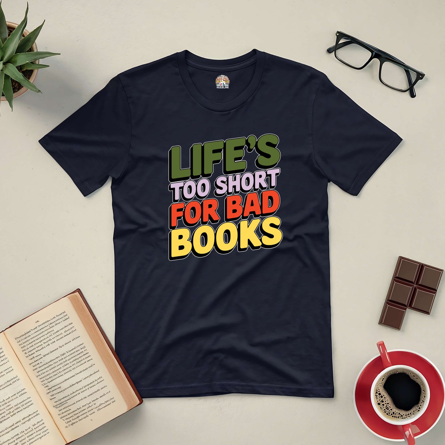 "Life's Too Short for Bad Books Tee with colorful text design, surrounded by books, glasses, chocolate, and a coffee cup on a table"