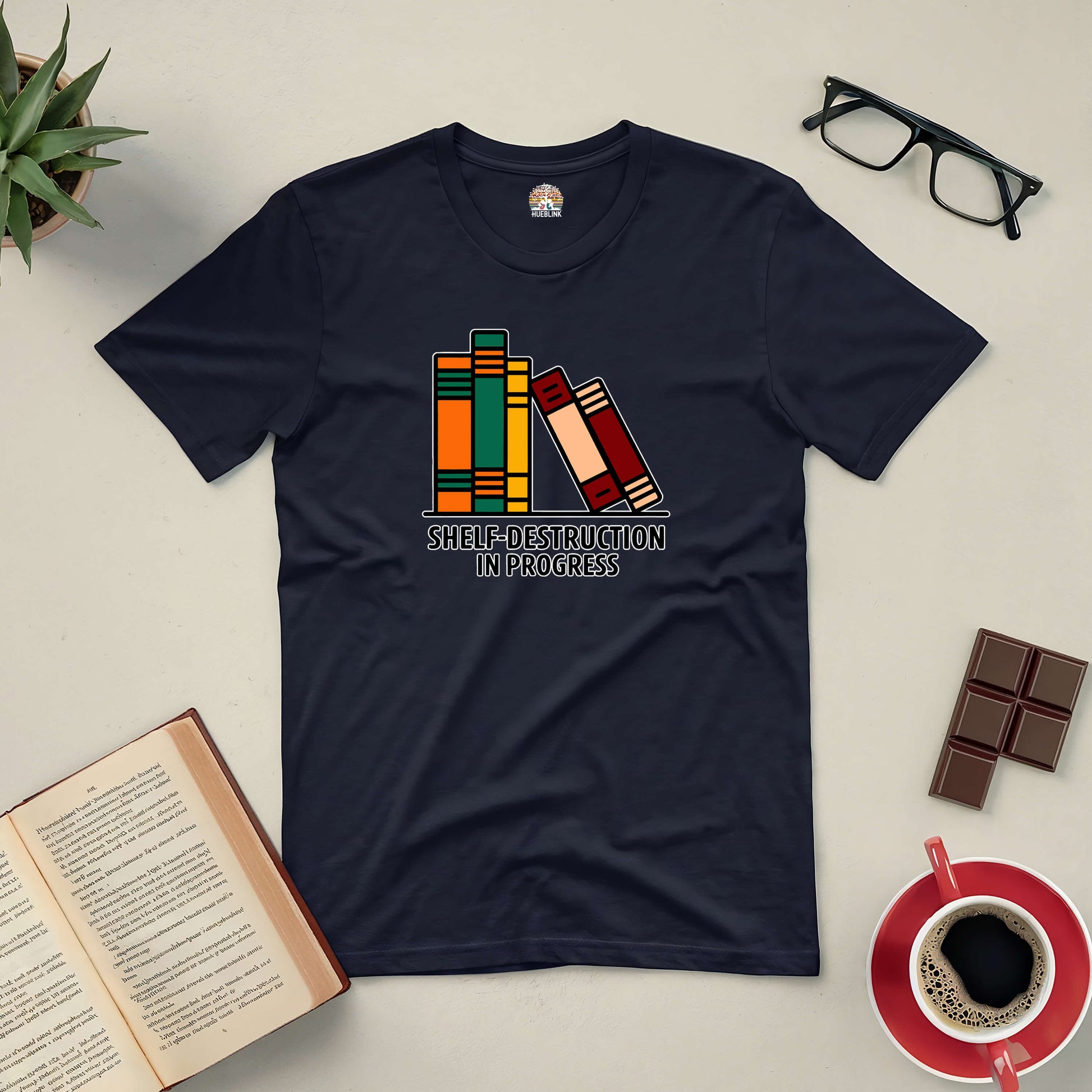 Black t-shirt featuring a graphic of leaning books with "Shelf-Destruction In Progress" text, surrounded by an open book, coffee, glasses, and chocolate.