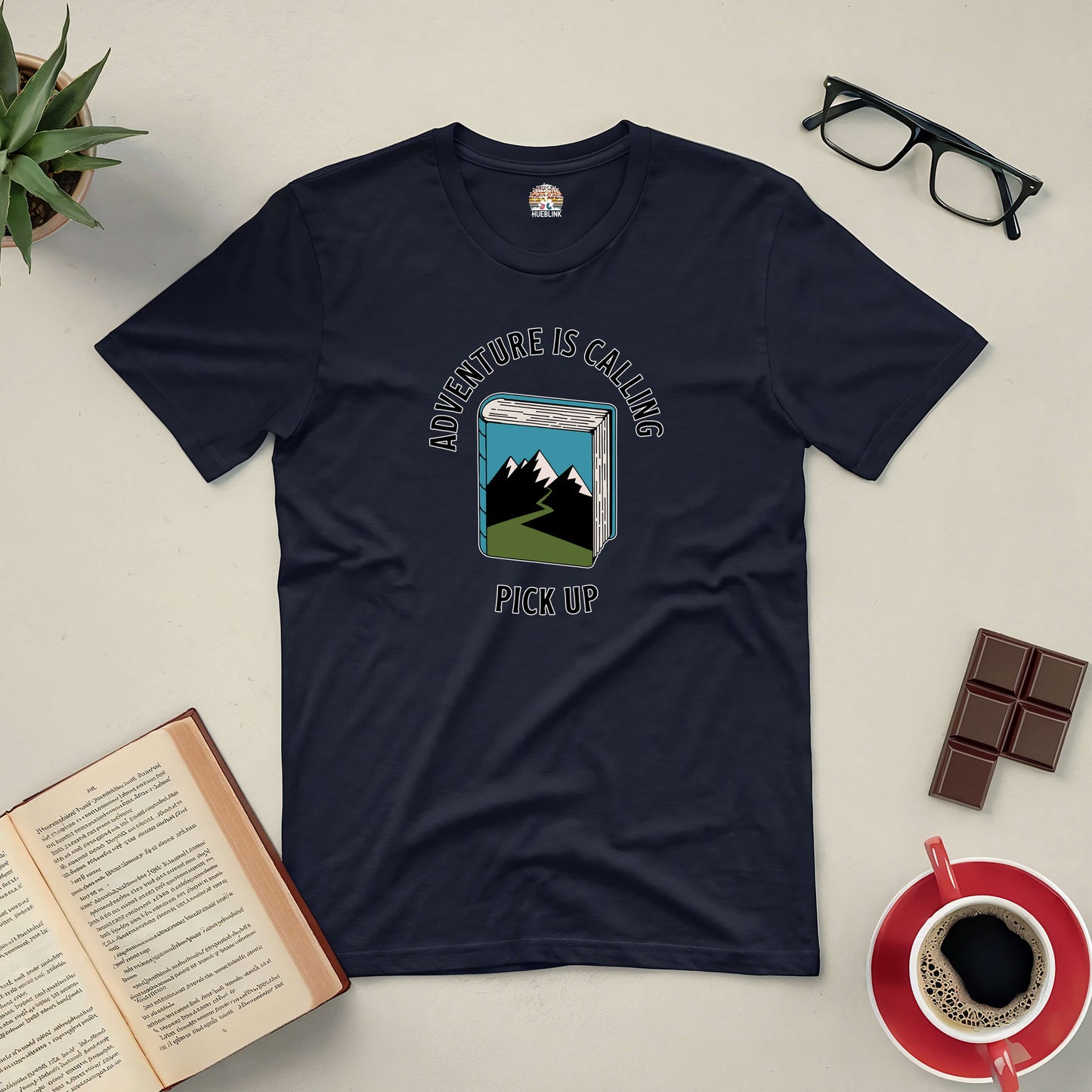 "Adventure Is Calling Pick Up Tee with mountain graphic surrounded by book, glasses, coffee, chocolate, and plant on a table"