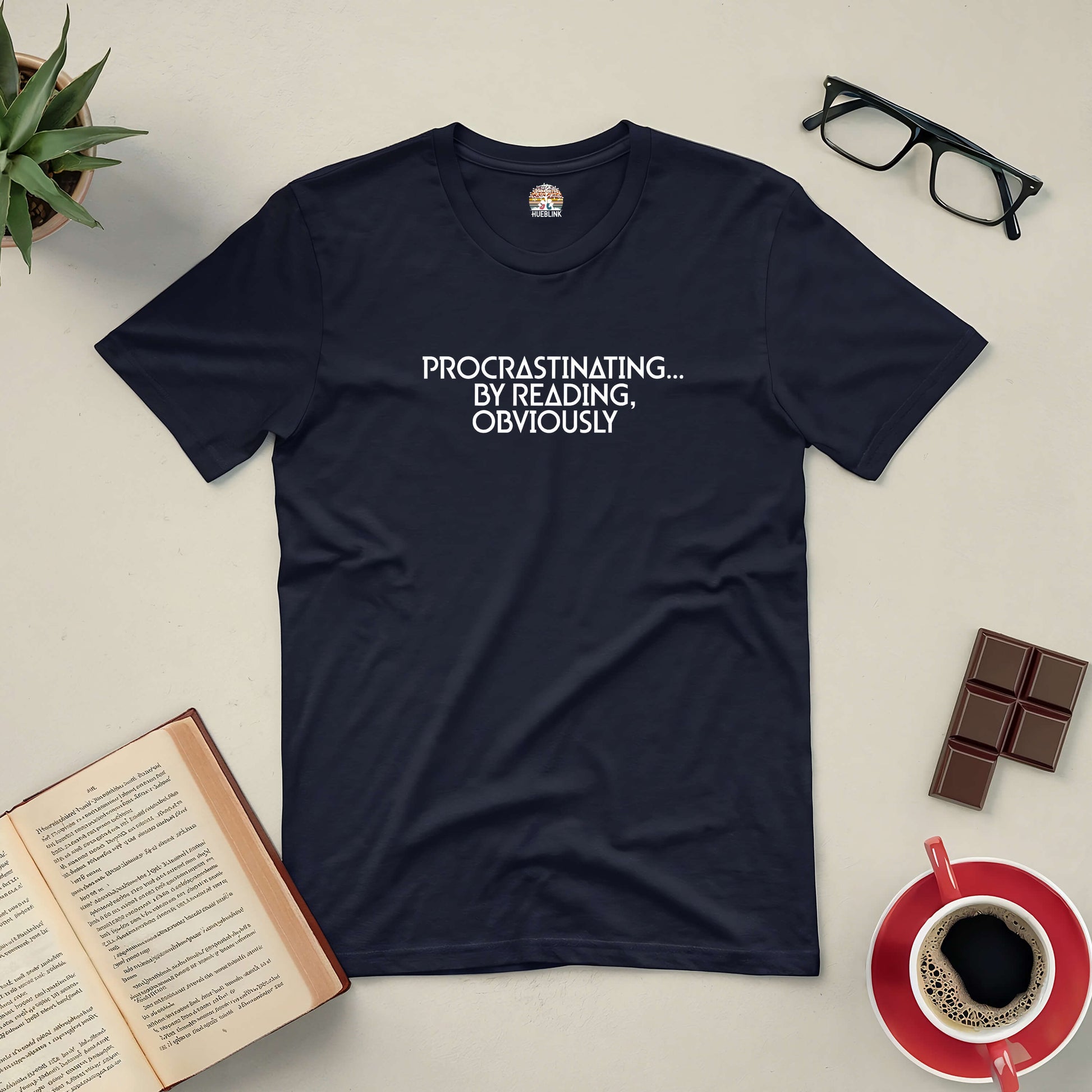 Black tee with "Procrastinating...By Reading Obviously" text, surrounded by a book, glasses, coffee, and chocolate on a table.