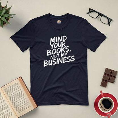 Black tee with "Mind Your Books, Not My Business" text, surrounded by a book, glasses, coffee, chocolates, and a plant.