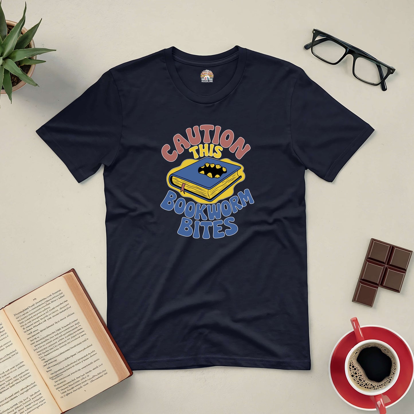 Black "Caution, This Bookworm Bites" tee with playful book design, surrounded by glasses, a book, chocolate, and coffee.