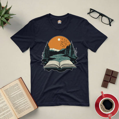 Black "Absorbed in Nature" tee with book and nature graphic, surrounded by a book, coffee, glasses, and plant on a flat surface.