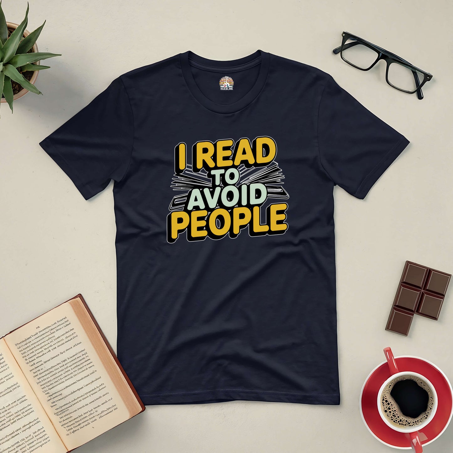 Black tee with "I Read To Avoid People" text, surrounded by open book, coffee, glasses, and chocolate, ideal for book lovers.