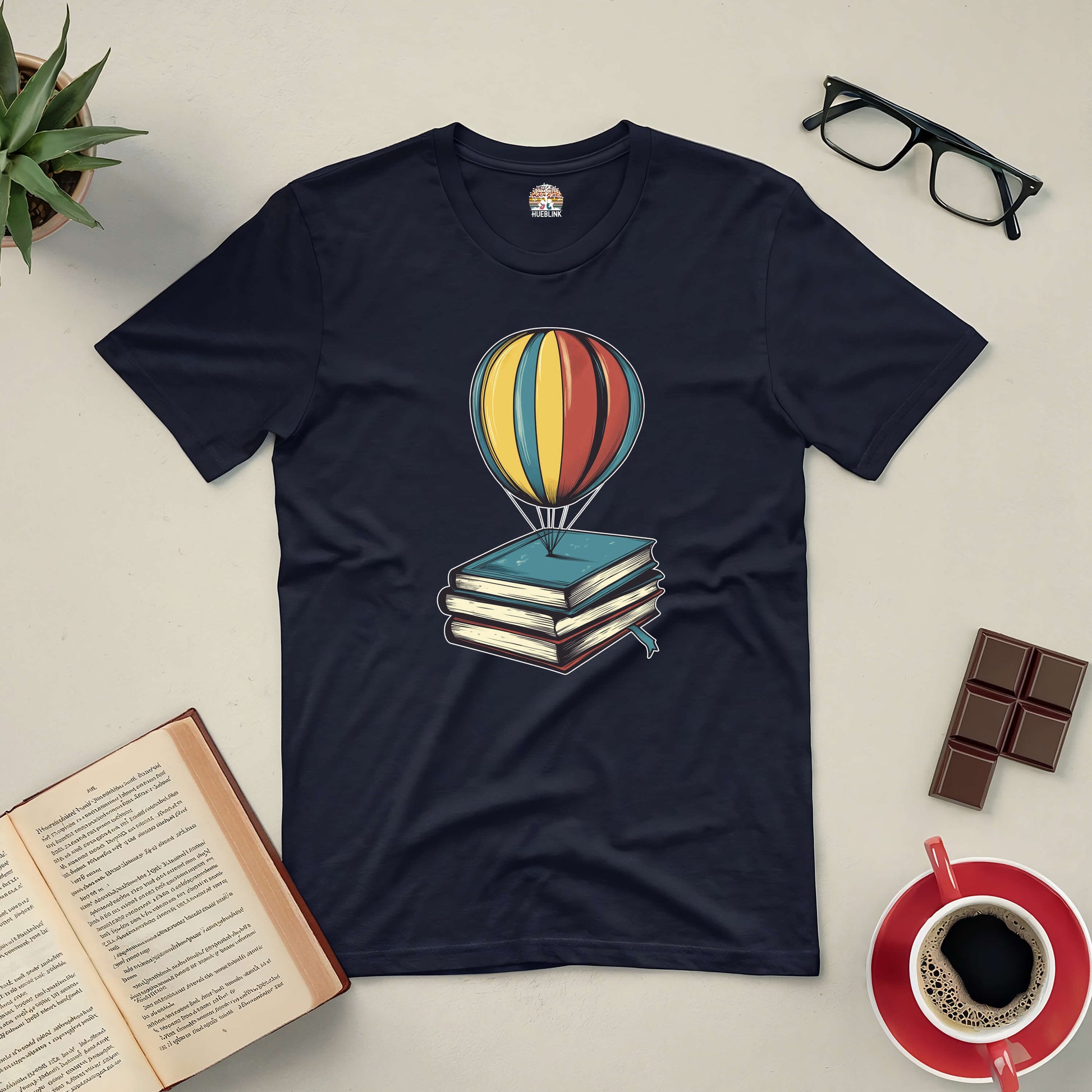 "Passport to Dreamland Tee with hot air balloon and books design, surrounded by coffee, chocolate, and reading essentials"