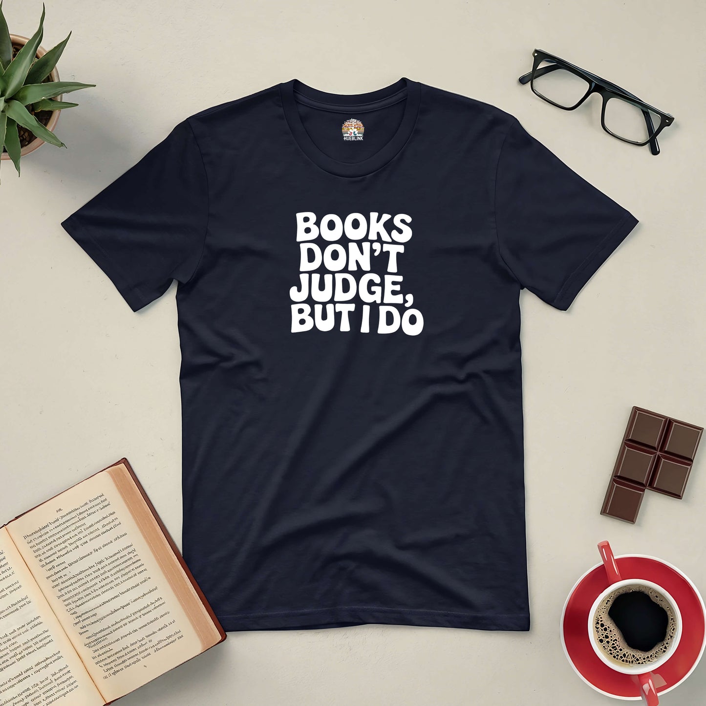 Black tee with "Books Don't Judge, But I Do" text, surrounded by an open book, glasses, coffee, chocolate, and a potted plant.