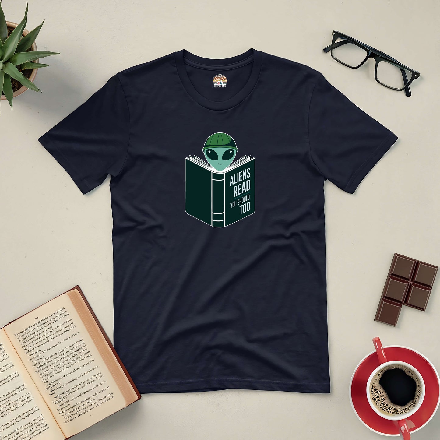 Intergalactic Habit Tee with alien reading design surrounded by an open book, glasses, and a coffee cup.