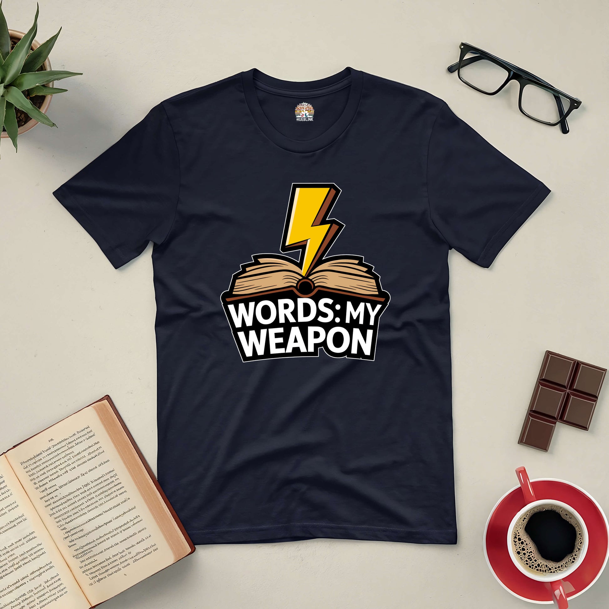 Black "Words: My Weapon" tee with book and lightning bolt design, displayed with open book, glasses, chocolate, and coffee.