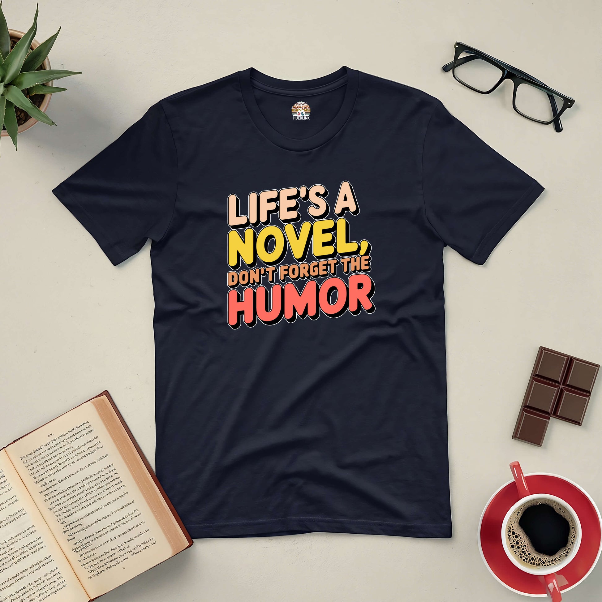 Black tee with "Life's a Novel, Don't Forget the Humor" text, surrounded by a book, glasses, chocolate, and coffee.