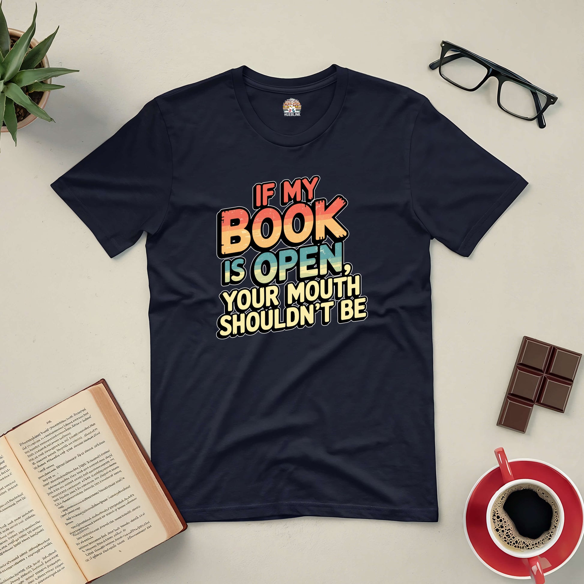 "Black tee with 'If My Book Is Open, Your Mouth Shouldn't Be' design for book lovers, shown with open book, glasses, chocolate, and coffee."