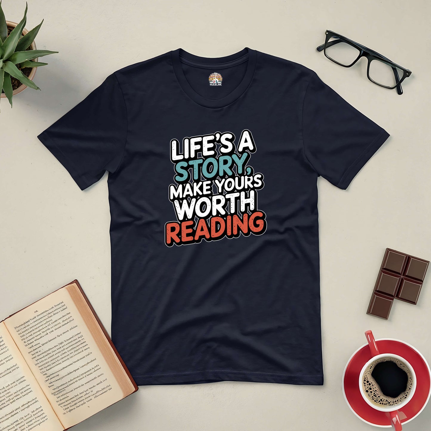 "Life's a Story Tee with motivational quote surrounded by a book, glasses, chocolate, plant, and coffee cup on a flat surface"