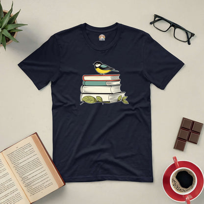 Black tee with a bird perched on stacked books, surrounded by glasses, chocolates, coffee, and an open book. Perfect for book lovers.