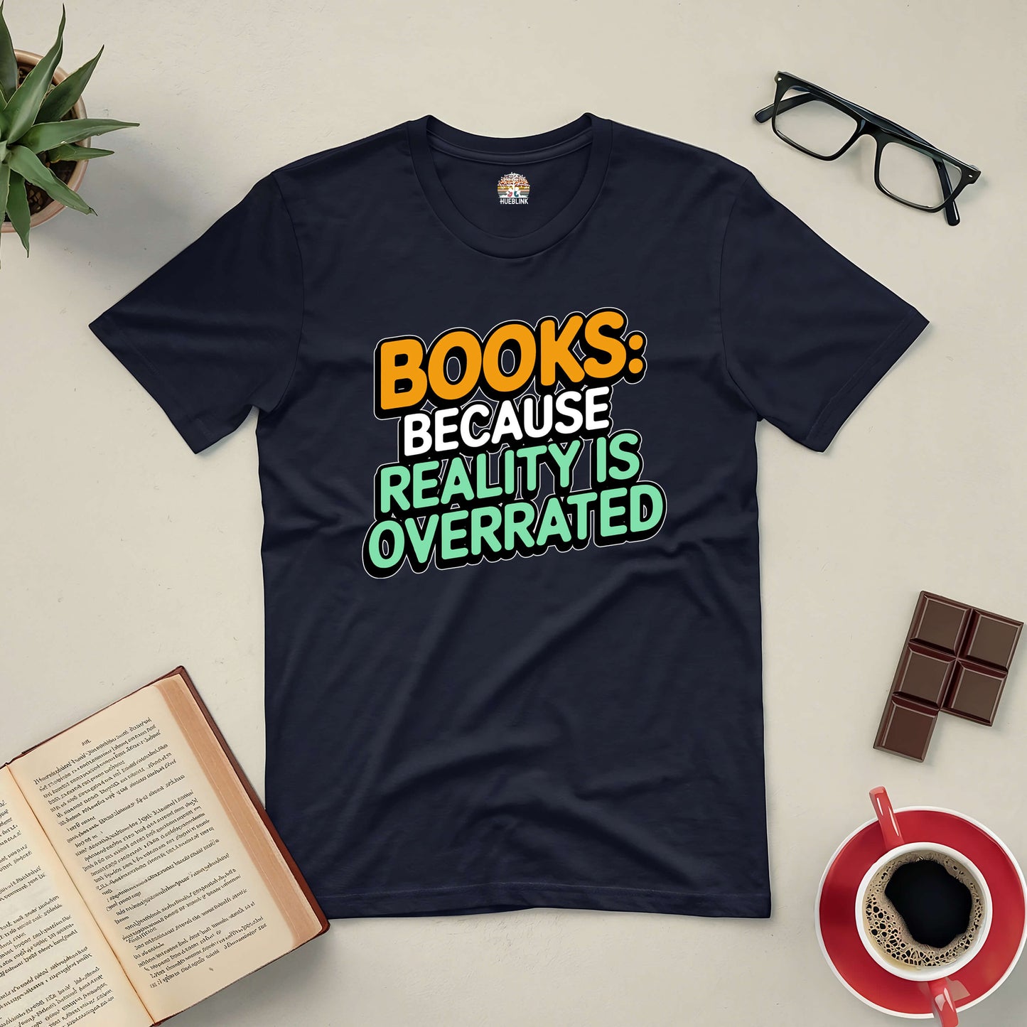 Black tee with "Books: Because Reality Is Overrated" text, surrounded by open book, glasses, chocolate, and coffee on a table.