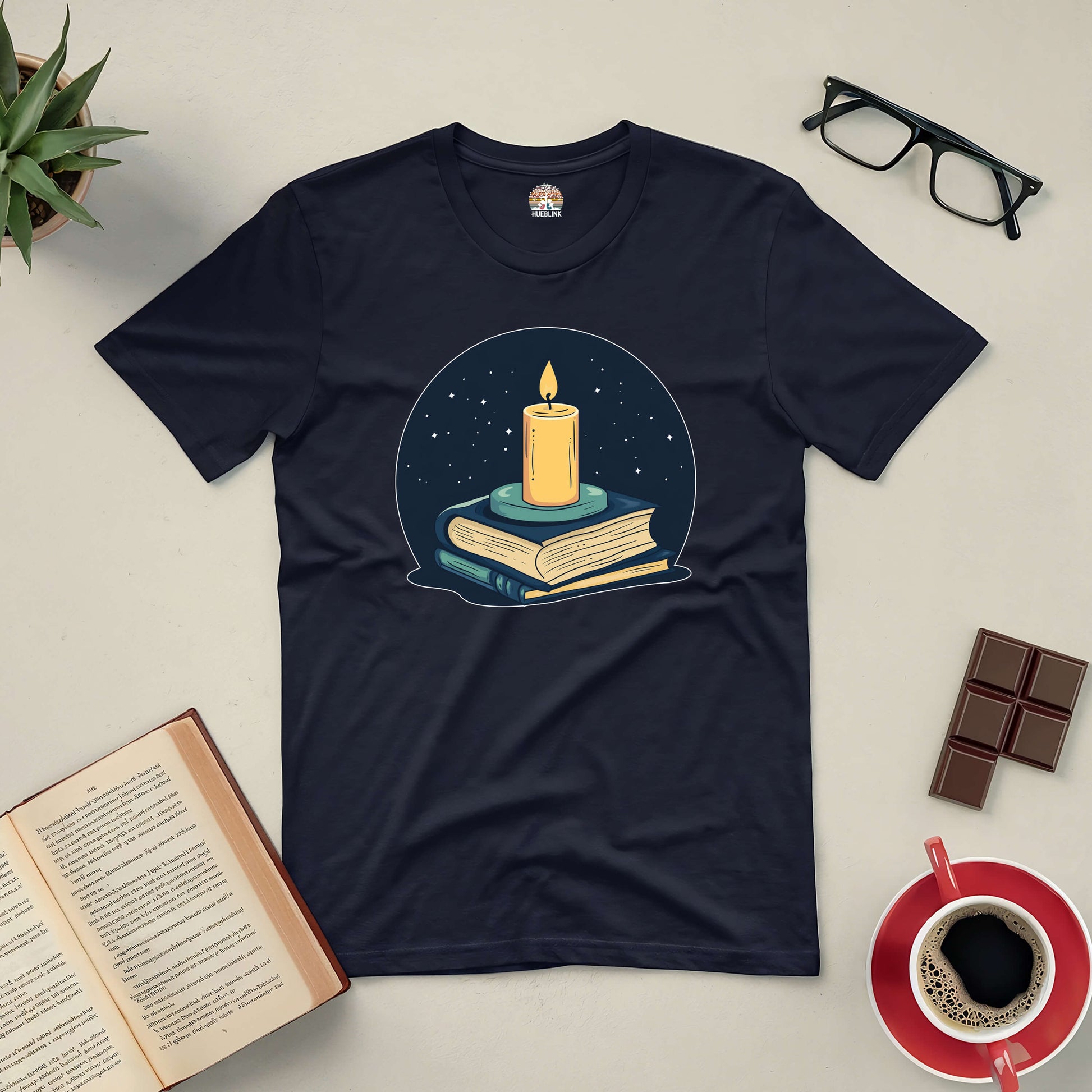 "Illuminate Your Mind Tee with candle on books design surrounded by reading essentials, coffee, and glasses"