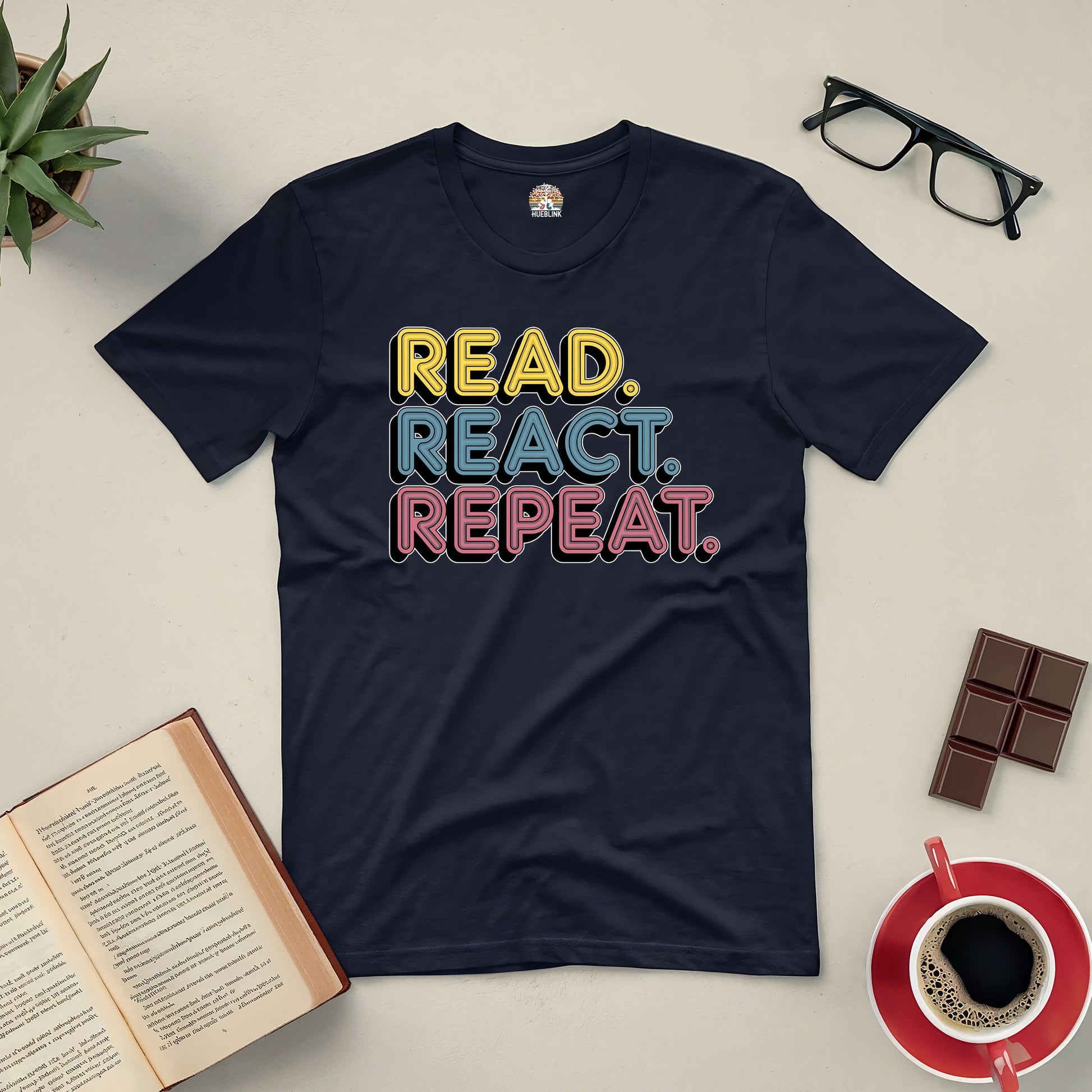 "Read React Repeat Tee with bold colorful text, surrounded by book, glasses, chocolate, and coffee on a table"