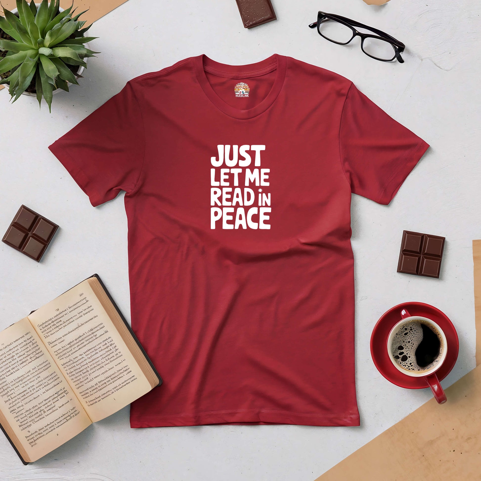 Red "Just Let Me Read in Peace" tee surrounded by a coffee cup, open book, eyeglasses, chocolate, and a plant on a table.