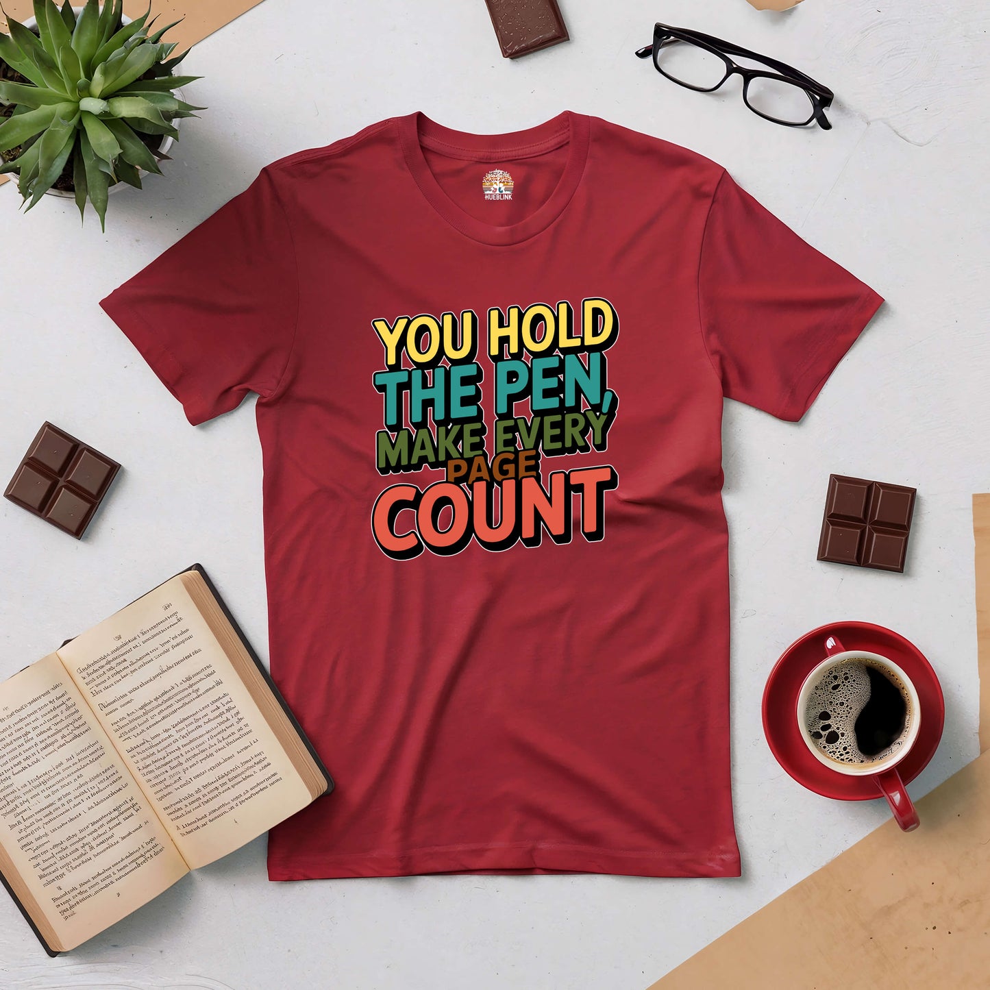 Red tee with "You Hold the Pen, Make Every Page Count" design, surrounded by books, glasses, and a coffee cup on a table.