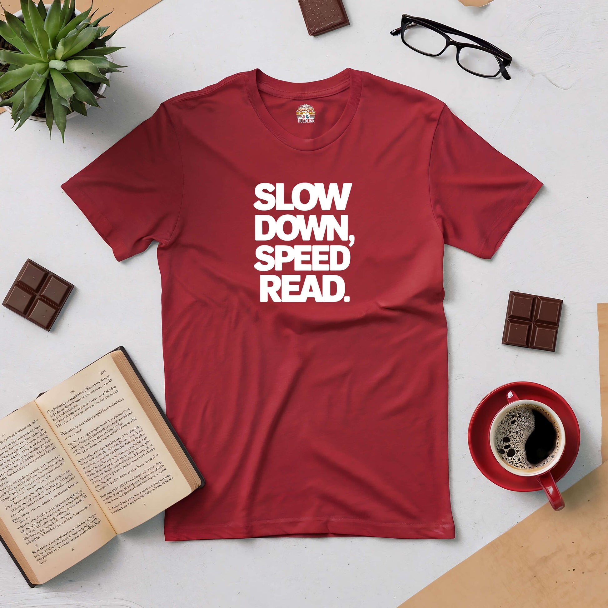 Red t-shirt with "Slow Down, Speed Read" design, surrounded by a cup of coffee, glasses, book, and chocolate on a table.