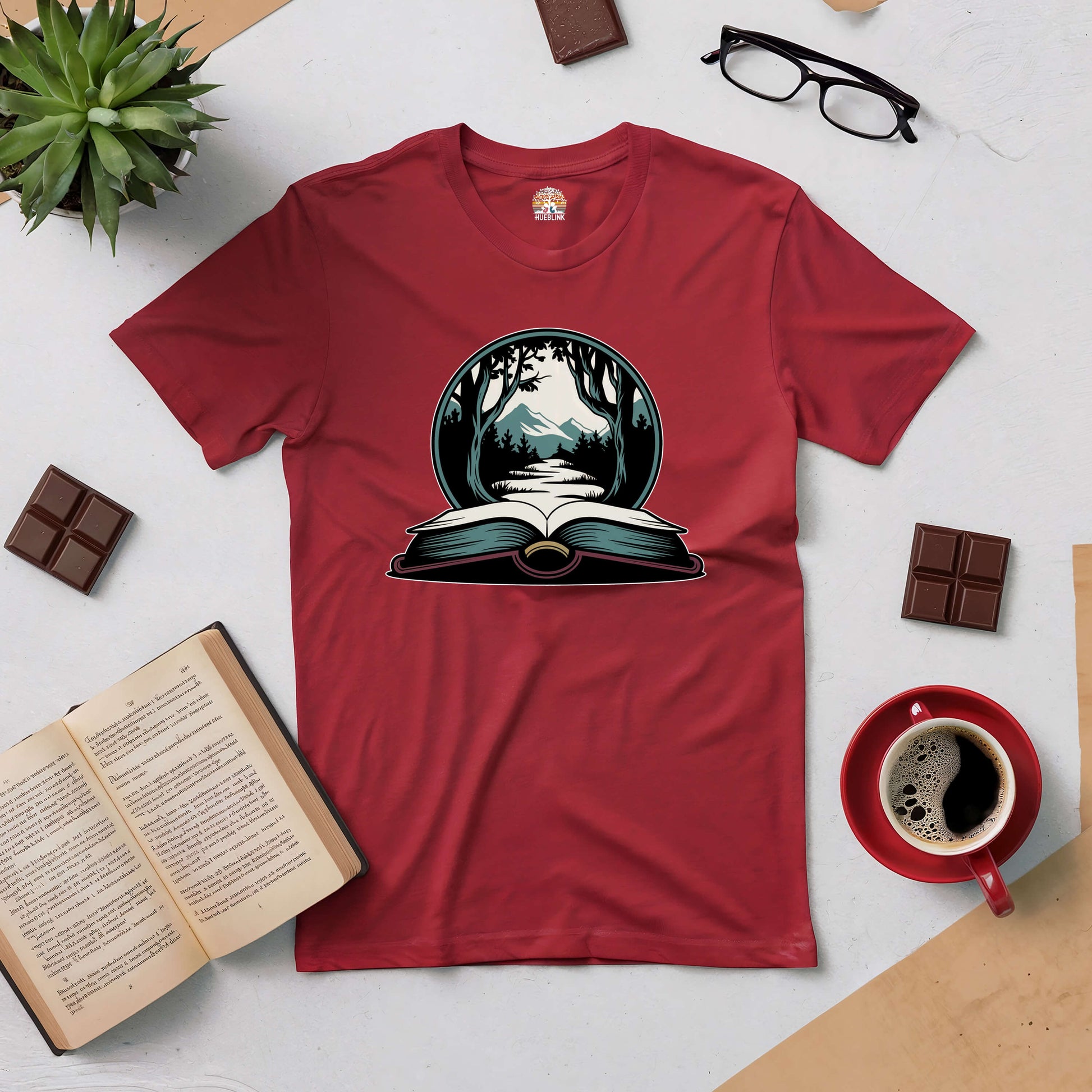 Red t-shirt featuring a wilderness design with an open book, surrounded by nature-inspired items like an open book, glasses, and coffee.