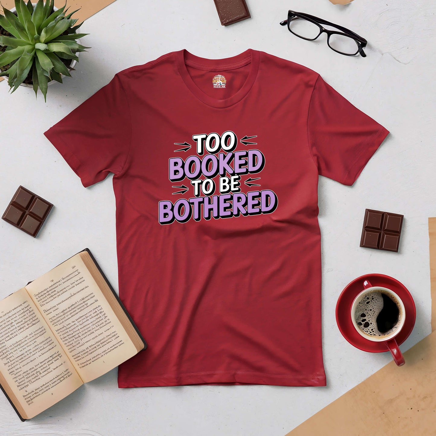 Red "Too Booked to Be Bothered" tee surrounded by a book, coffee, chocolate, and glasses, perfect for avid readers.