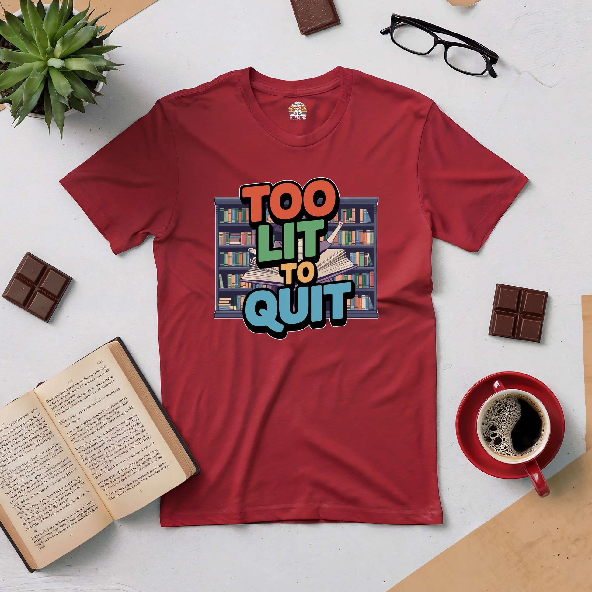 Red "Too Lit To Quit" tee surrounded by books, chocolate, coffee, and glasses on a light background. Perfect for book lovers.