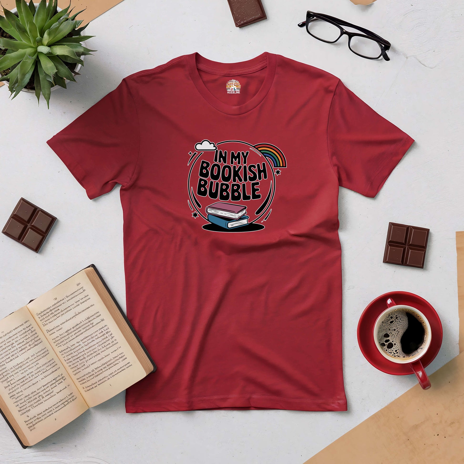 Red "In My Bookish Bubble" tee surrounded by an open book, coffee cup, glasses, chocolates, and a small plant, ideal for book lovers.