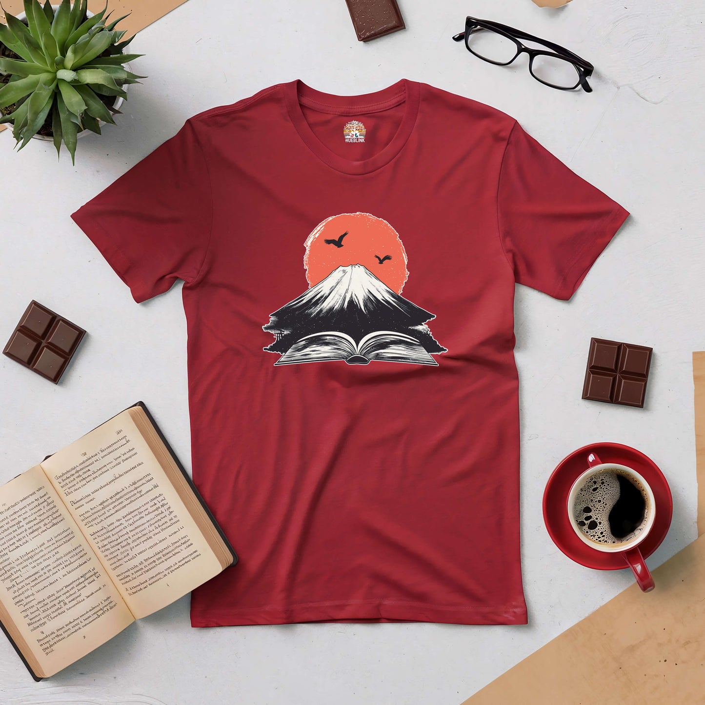 Red Eruption of Imagination Tee featuring volcano and book design, surrounded by coffee, chocolate, open book, and glasses.