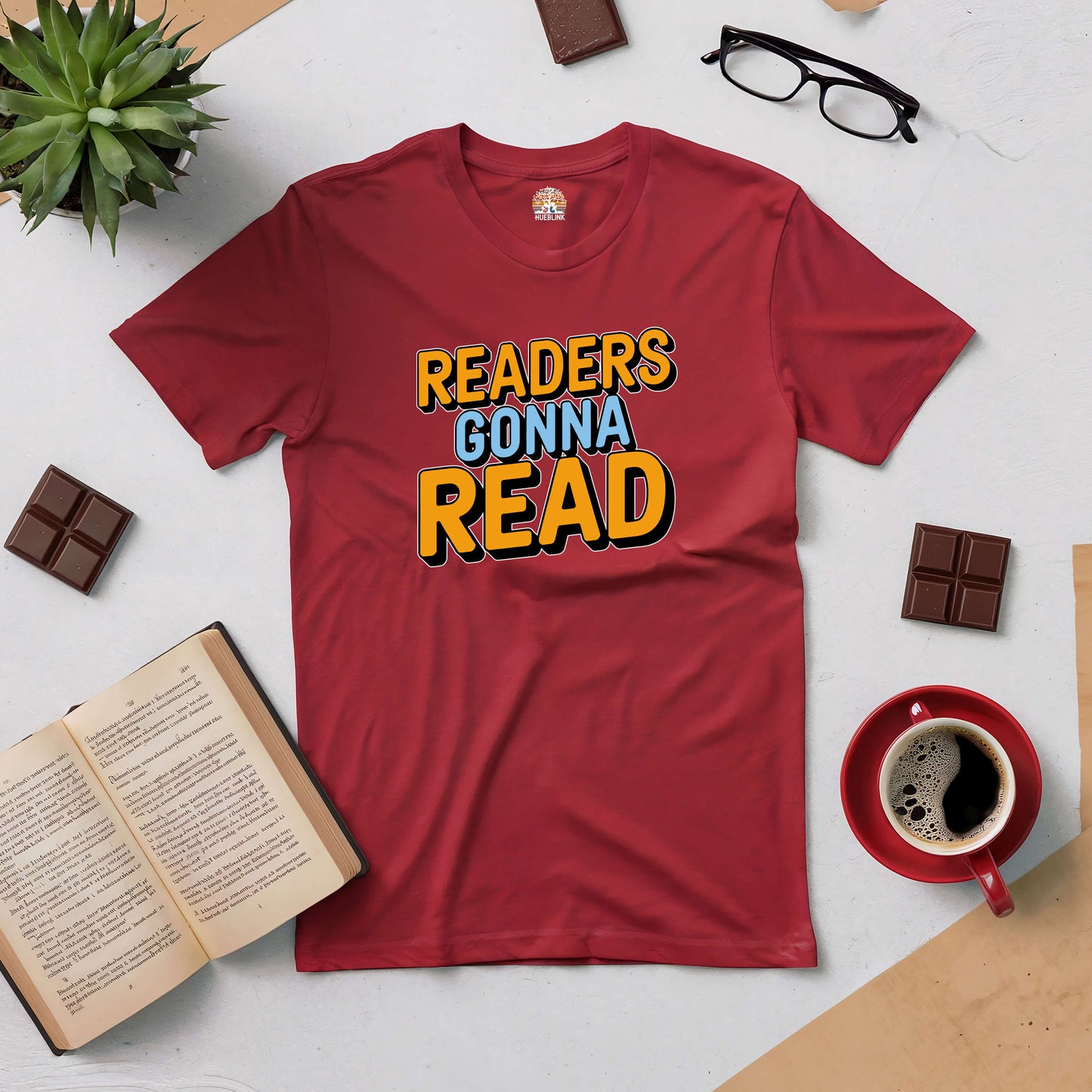 Red "Readers Gonna Read" tee surrounded by coffee, glasses, chocolate, and an open book on a white surface.