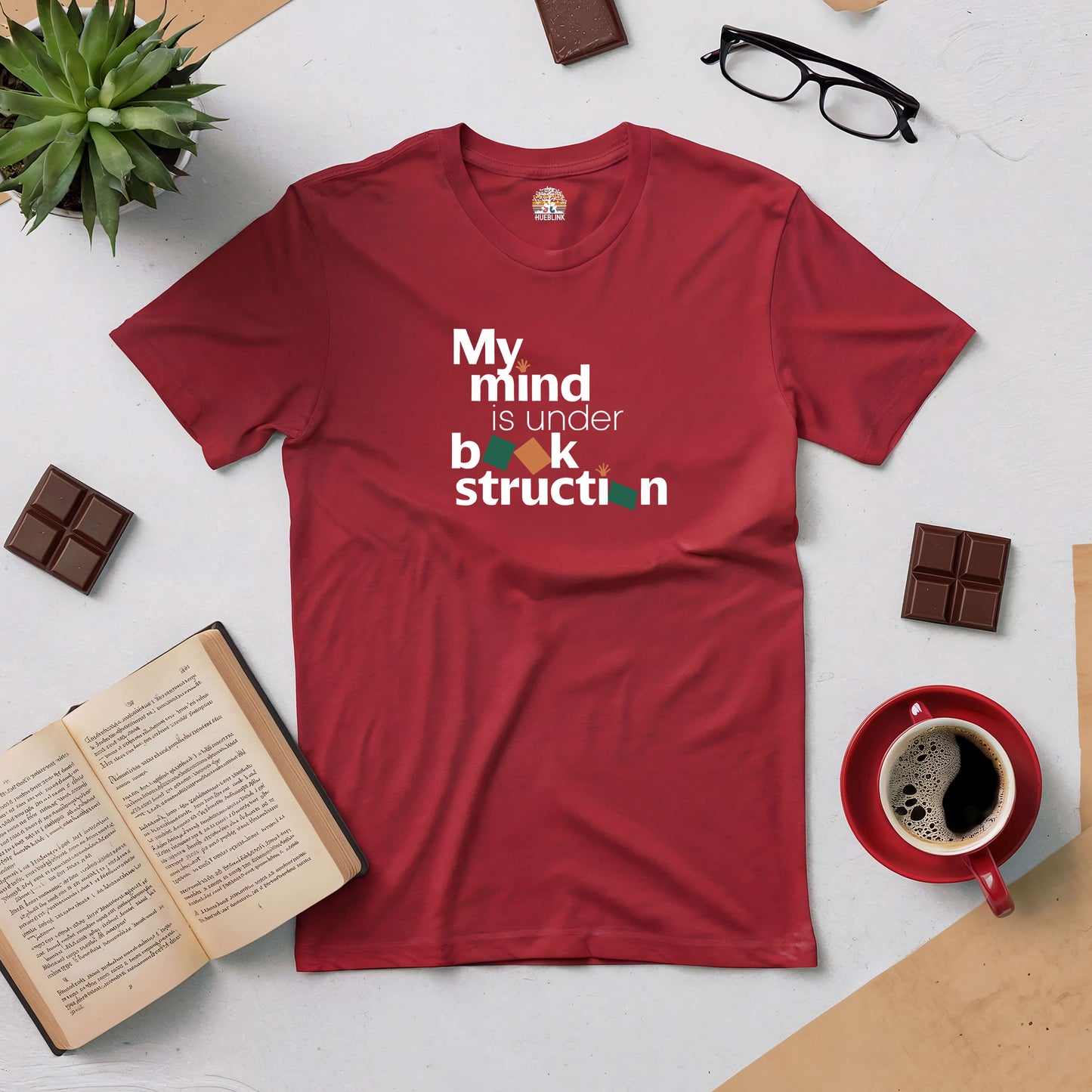 Red "My Mind is Under Book-struction" T-shirt on a flat lay with books, coffee, plant, chocolates, and glasses.