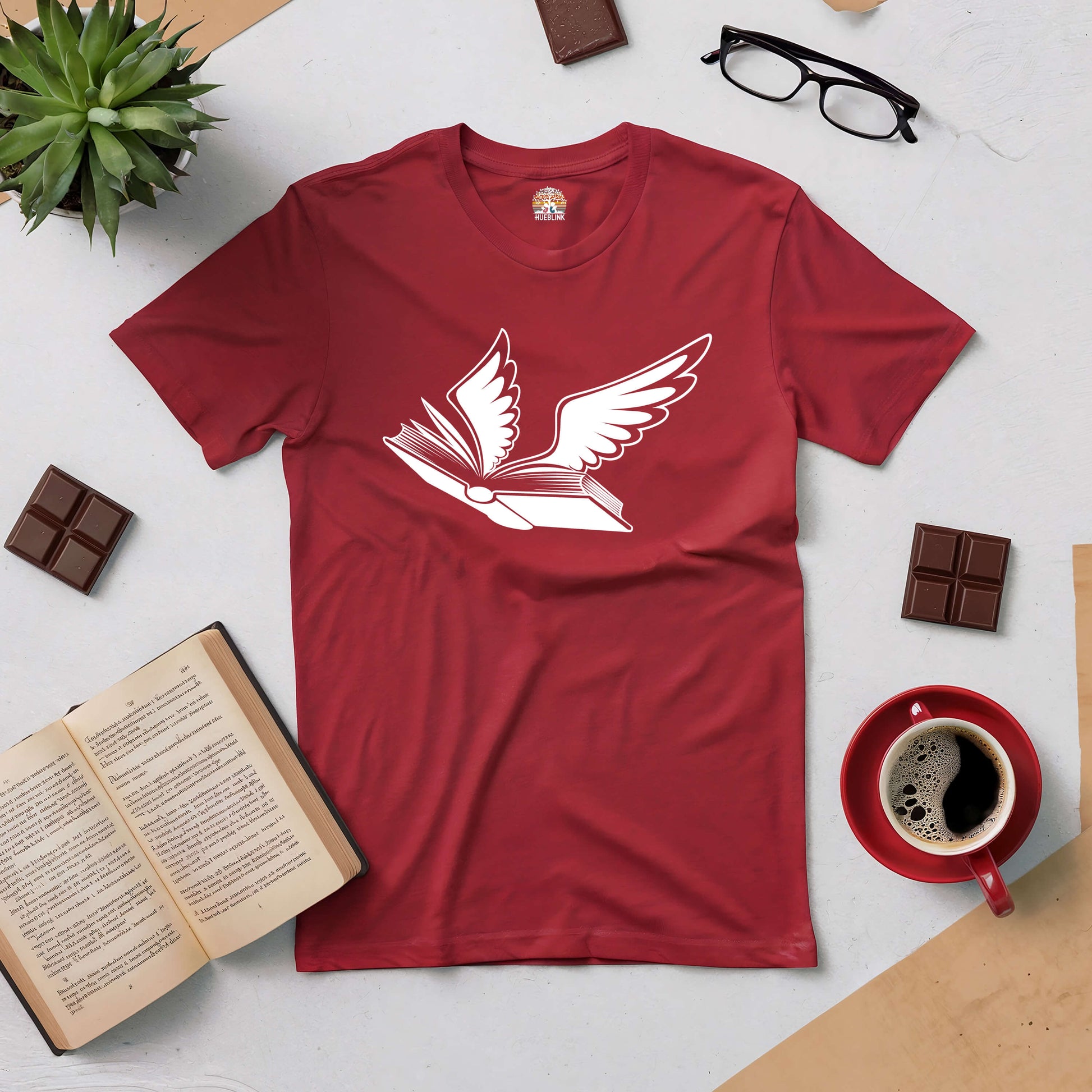 Red Bird of Hermes Tee with winged book design, surrounded by coffee, chocolate, glasses, open book, and plant on a table.