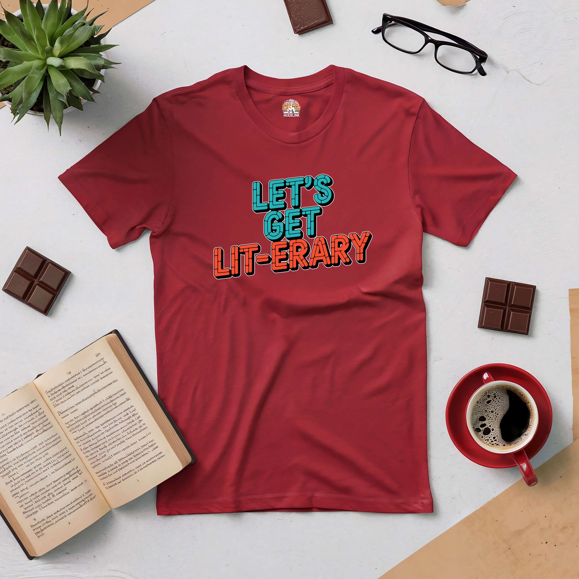 Red "Let's Get Lit-erary" tee surrounded by books, glasses, and a coffee, ideal for lively book lovers.