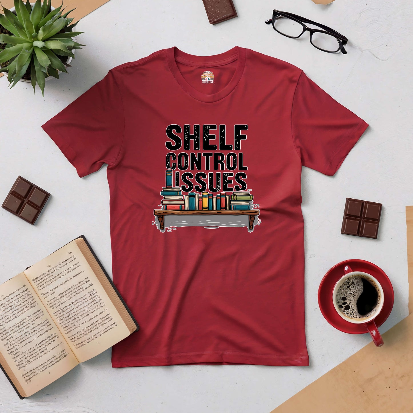 Red "Shelf Control Issues" tee with bookshelf design, surrounded by books, coffee, glasses, and chocolate on a table.