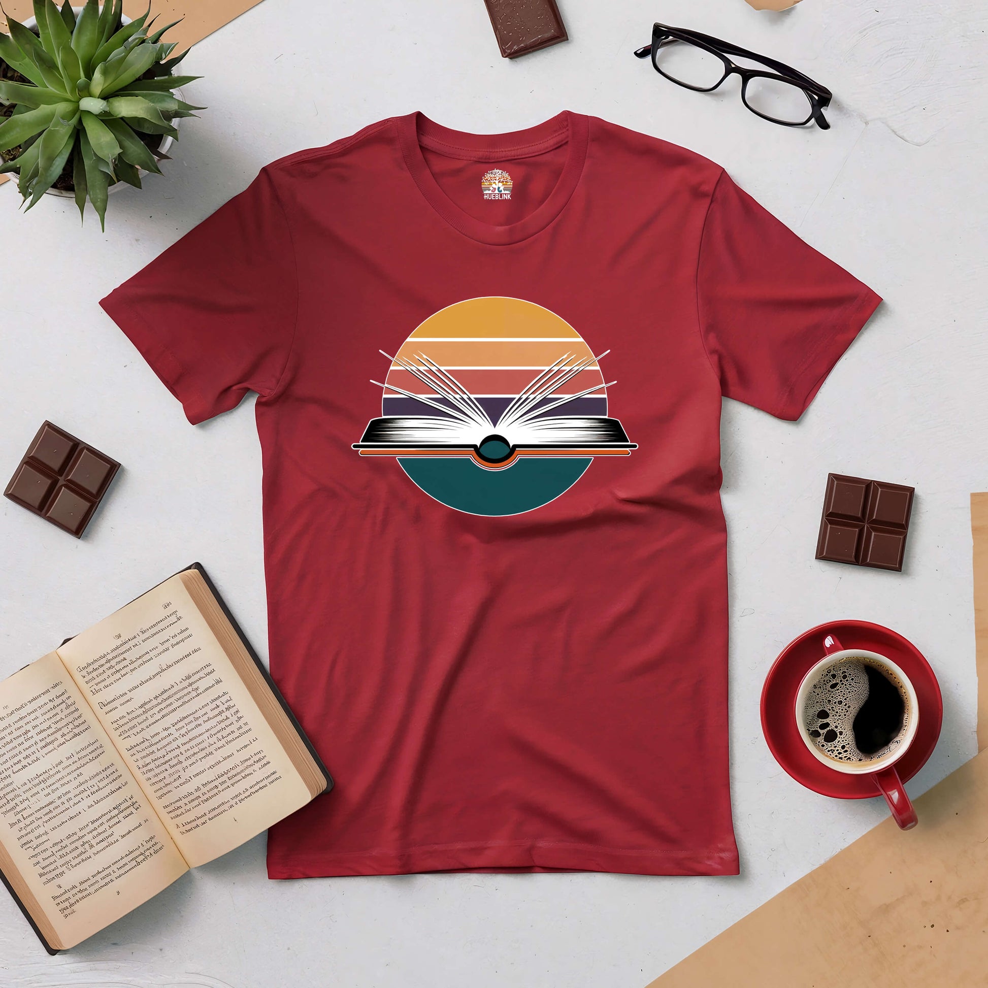 "Gateway to New Horizons Tee with book design, surrounded by an open book, plant, glasses, chocolate, and coffee on a table."