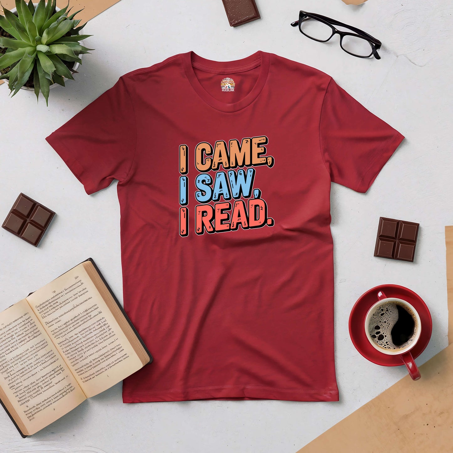 Red tee with "I Came, I Saw, I Read" text, surrounded by books, coffee, chocolate, glasses, and plant on a flatlay.