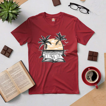 Red Beachside Bibliophile tee with sunset and book graphic, surrounded by coffee, open book, glasses, plant, and chocolate.