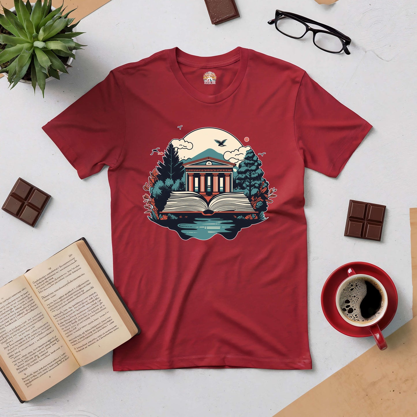 "Gateway to Knowledge Tee with library and nature design, symbolizing wisdom through books, laid out with plants, chocolate, and coffee"