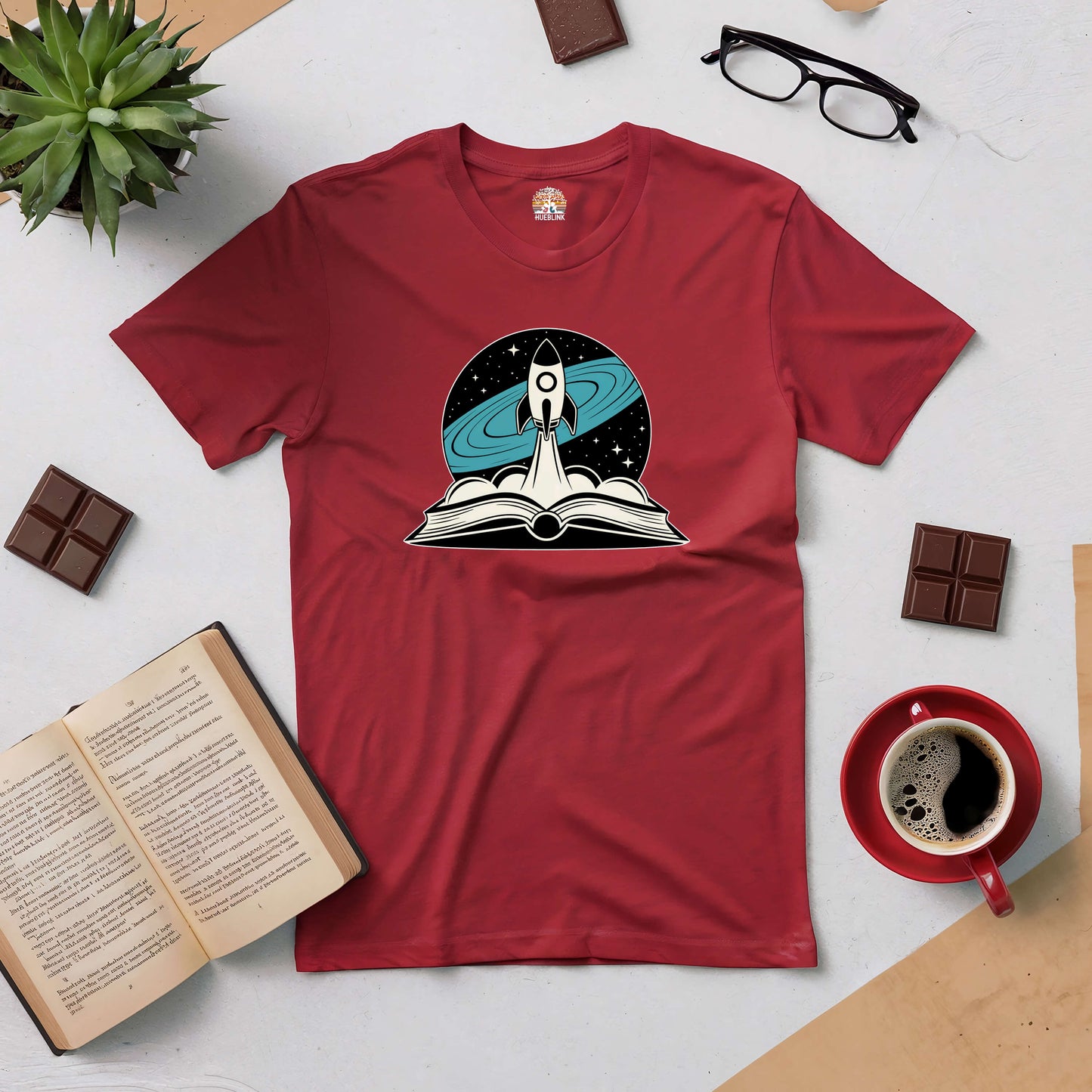 Red tee with rocket launching from an open book, surrounded by glasses, coffee, chocolate, and a plant, symbolizing adventure in reading.