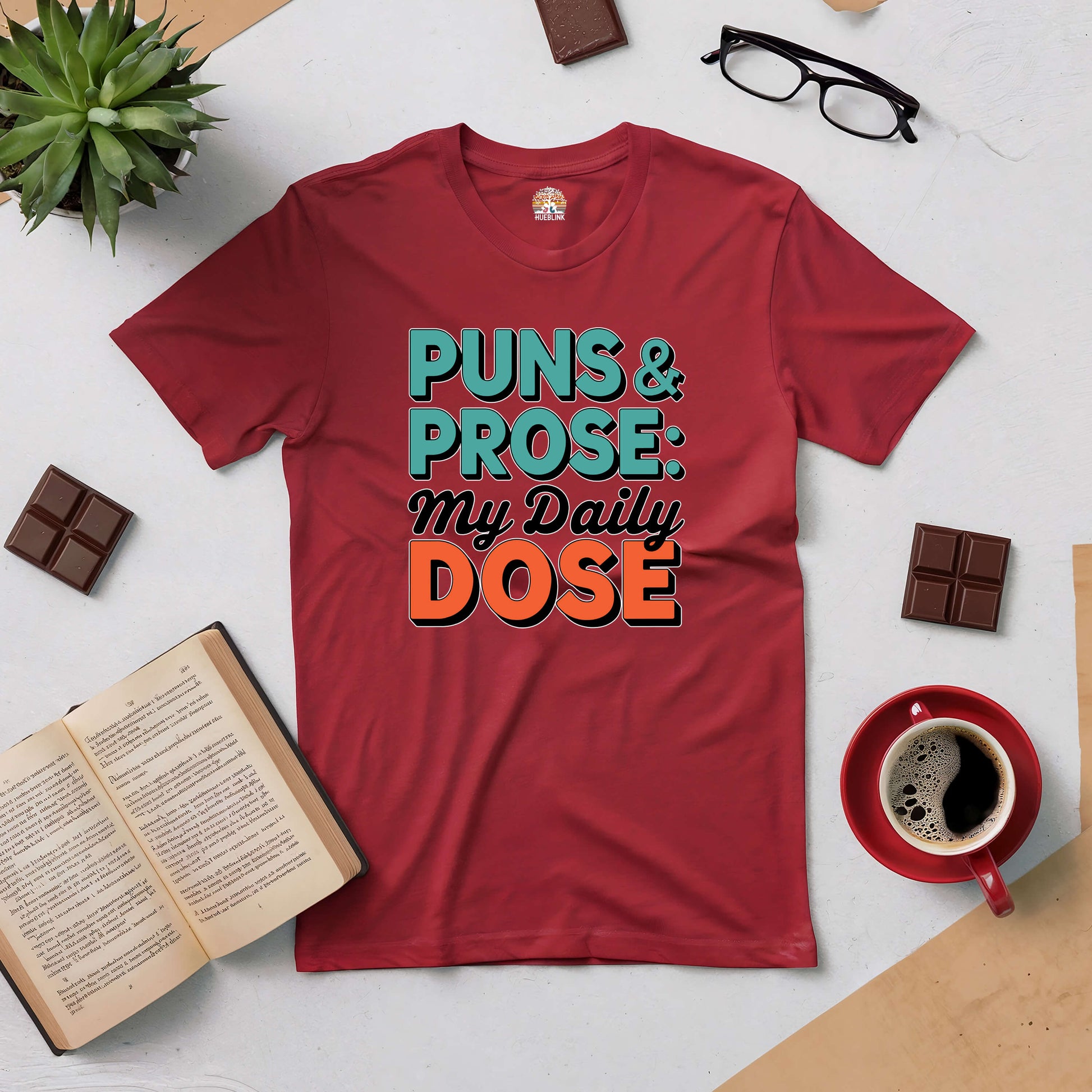 Red t-shirt with "Puns & Prose: My Daily Dose" text, surrounded by books, glasses, coffee, and chocolate on a creative flat lay.