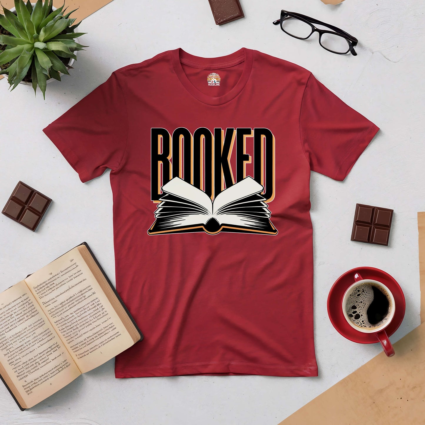 "Red Booked Tee with open book graphic, surrounded by books, coffee, glasses, and chocolate on a table, ideal for book lovers."