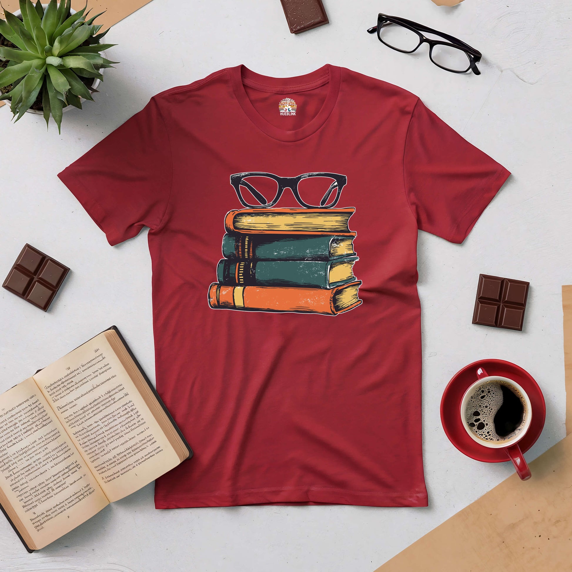 Red t-shirt with vintage books and glasses design, styled with coffee, open book, chocolates, and green plant for readers and fashion lovers.