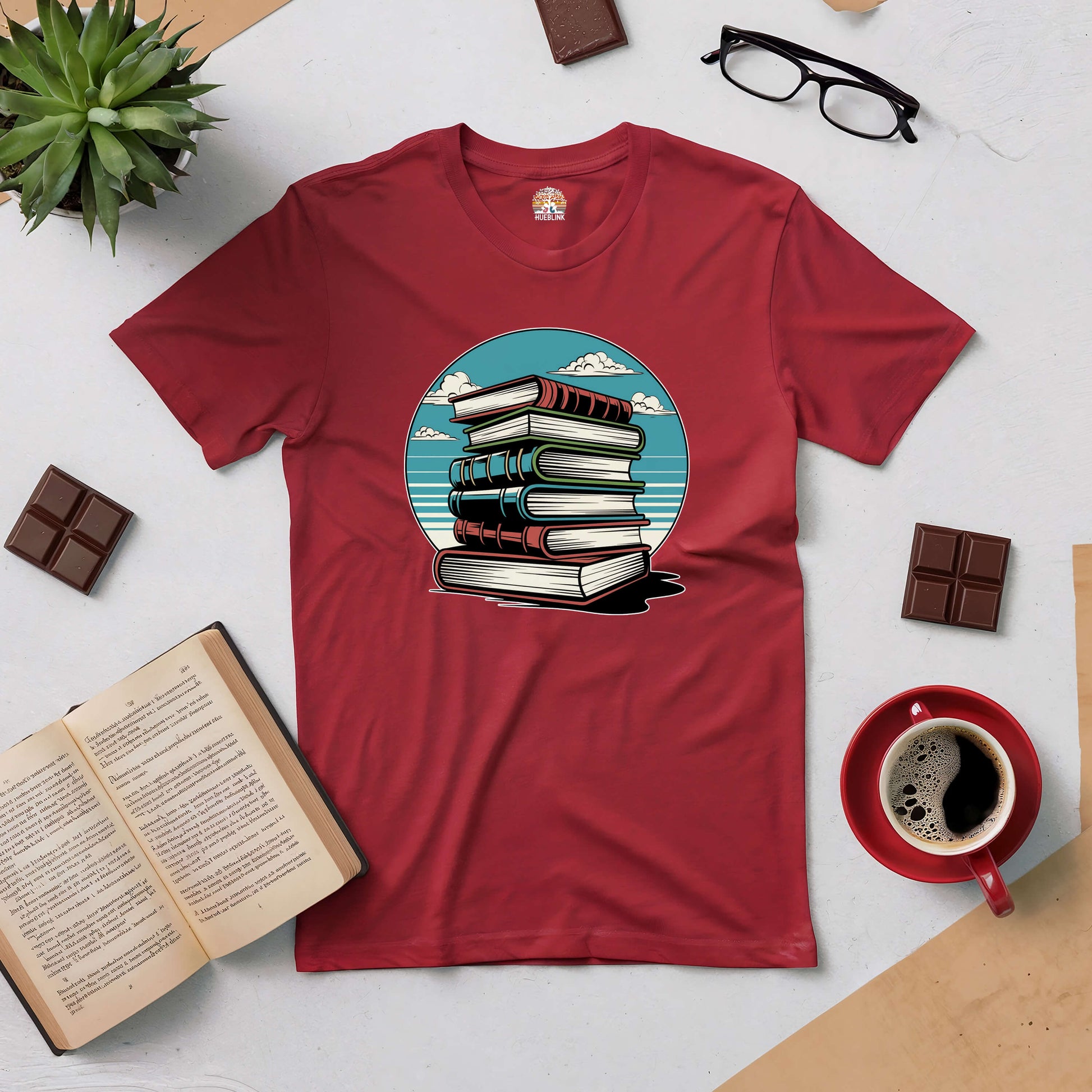 Red Silent Knowledge Tee featuring a stack of books under an open sky, surrounded by reading accessories like coffee and chocolates.