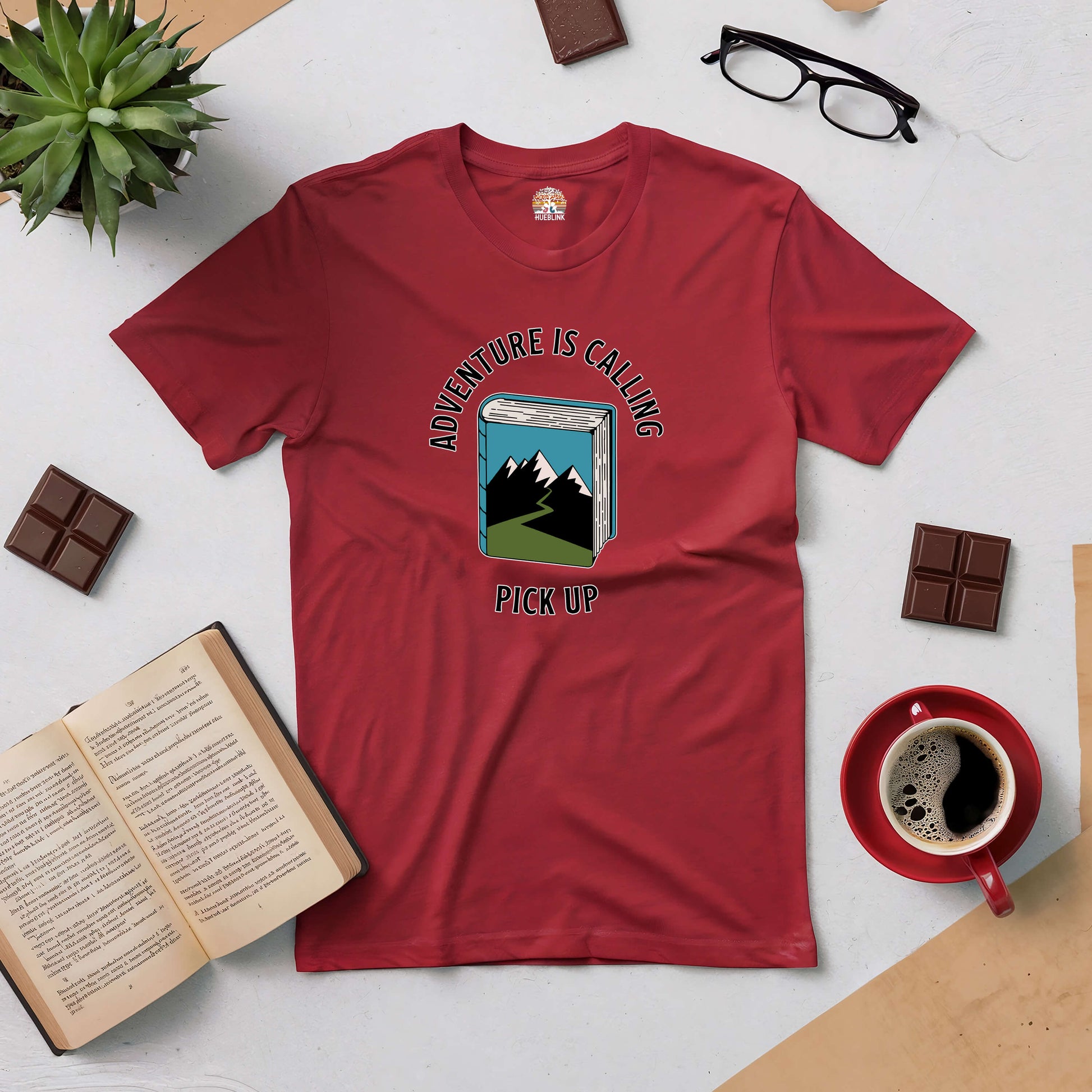 Red "Adventure Is Calling, Pick Up" t-shirt with book and mountain design, perfect for explorers, surrounded by coffee, books, and chocolate.